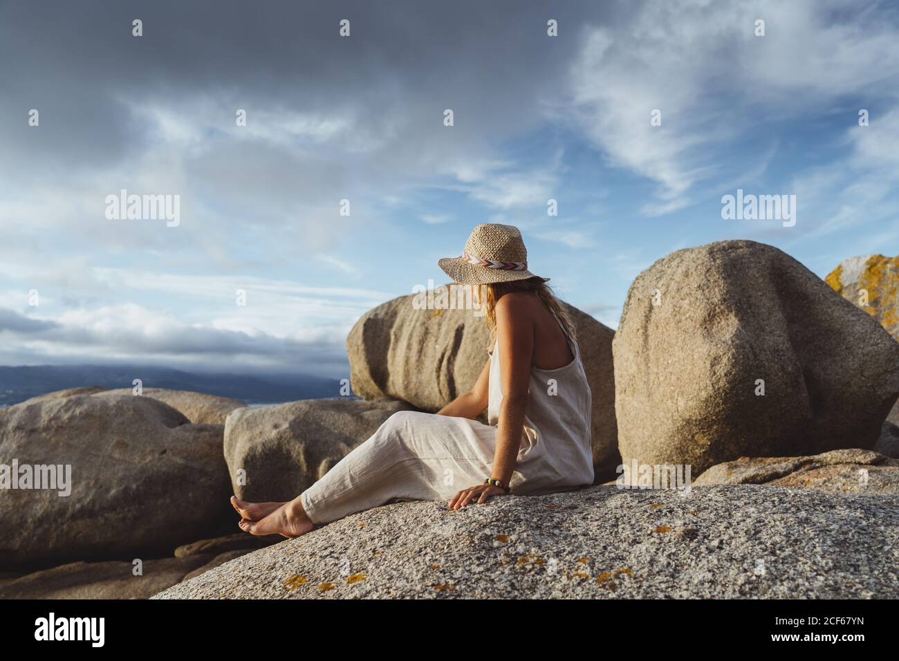Seitenansicht einer glücklichen und optimistischen Dame im Strohhut Sitzen vor den Klippen am Meer und lächeln weg in Abendlicht Stockfoto