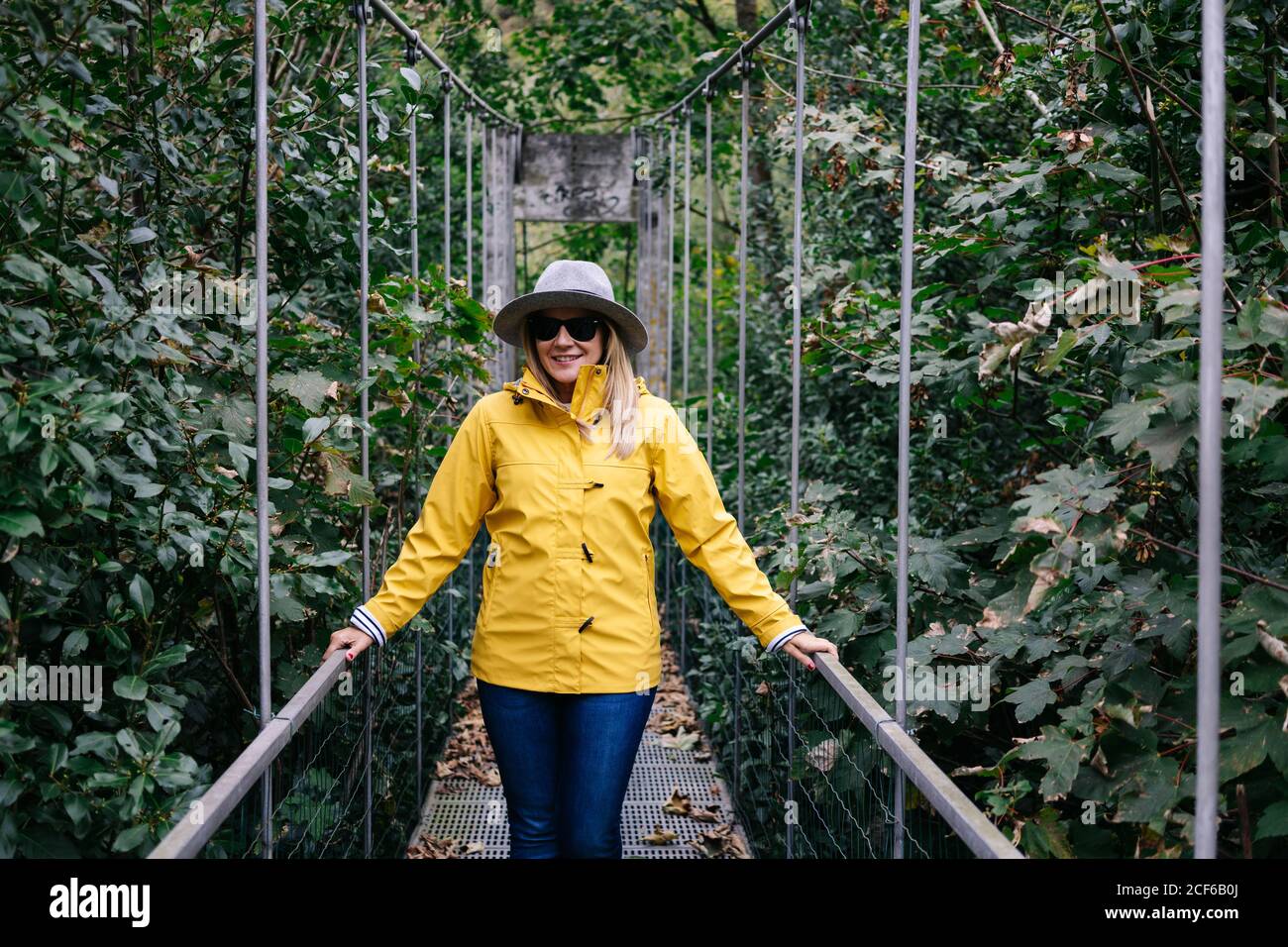 Glückliche Frau in Hut und Sonnenbrille in gelber Jacke, die im Sommer auf der Hängebrücke steht und den Urlaub genießt Stockfoto