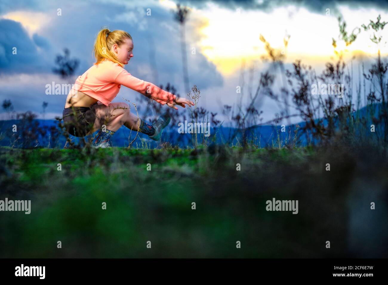 Seitenansicht einer starken Frau, die beim funktionellen Training auf dem Spielfeld mit einem Bein gegen den wolkigen Sonnenuntergang hockt Stockfoto