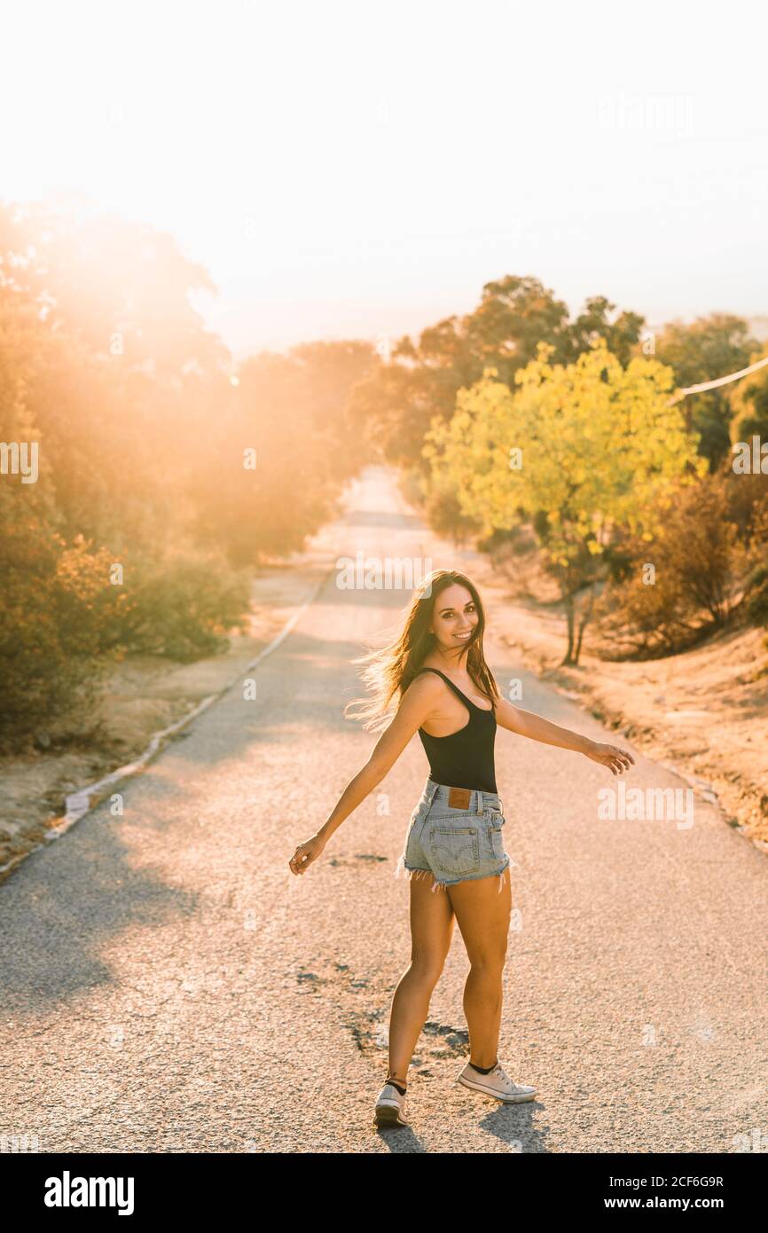 Rückansicht des Inhalts schöne Frau in jean-Shorts, die über die Schulter blickt, während sie auf einer landschaftlich reizvollen Straße mit Hintergrundbeleuchtung spazieren geht Stockfoto