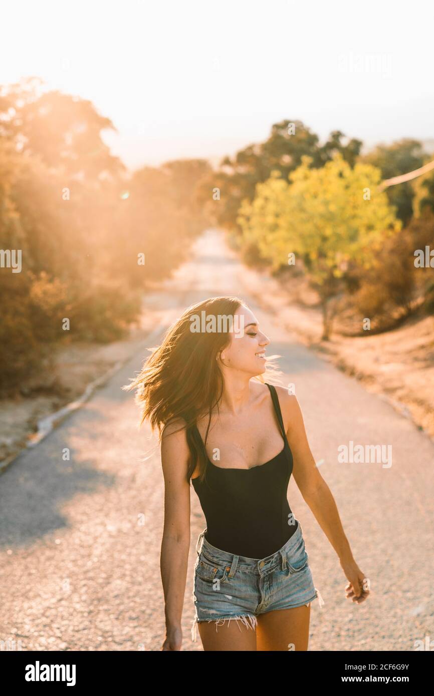 Rückansicht des Inhalts schöne Frau in jean-Shorts, die über die Schulter blickt, während sie auf einer landschaftlich reizvollen Straße mit Hintergrundbeleuchtung spazieren geht Stockfoto