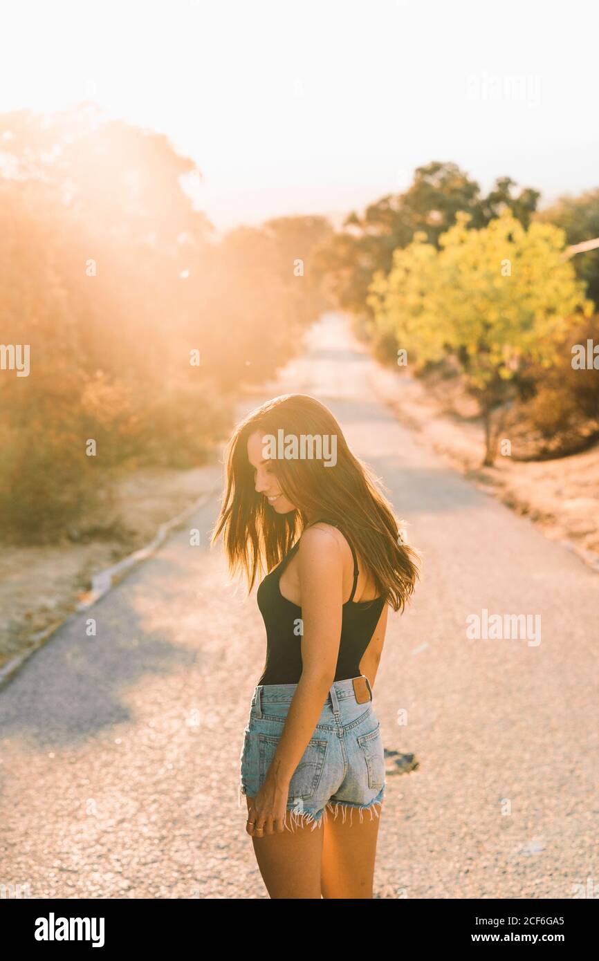 Rückansicht des Inhalts schöne Frau in jean-Shorts, die über die Schulter blickt, während sie auf einer landschaftlich reizvollen Straße mit Hintergrundbeleuchtung spazieren geht Stockfoto