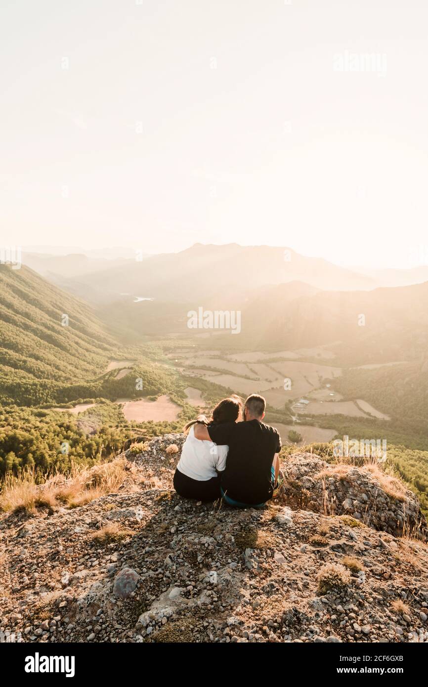 Rückansicht von romantischen paar Touristen in legerer Kleidung Sitzen auf Steinkante der Klippe umarmen und genießen malerisch Landschaft bei sonnigem Tag Stockfoto