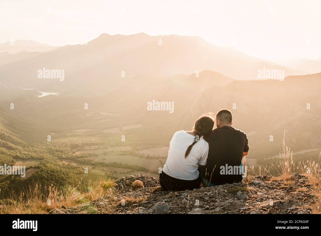 Rückansicht von romantischen paar Touristen in legerer Kleidung Sitzen auf Steinkante der Klippe umarmen und genießen malerisch Landschaft bei sonnigem Tag Stockfoto