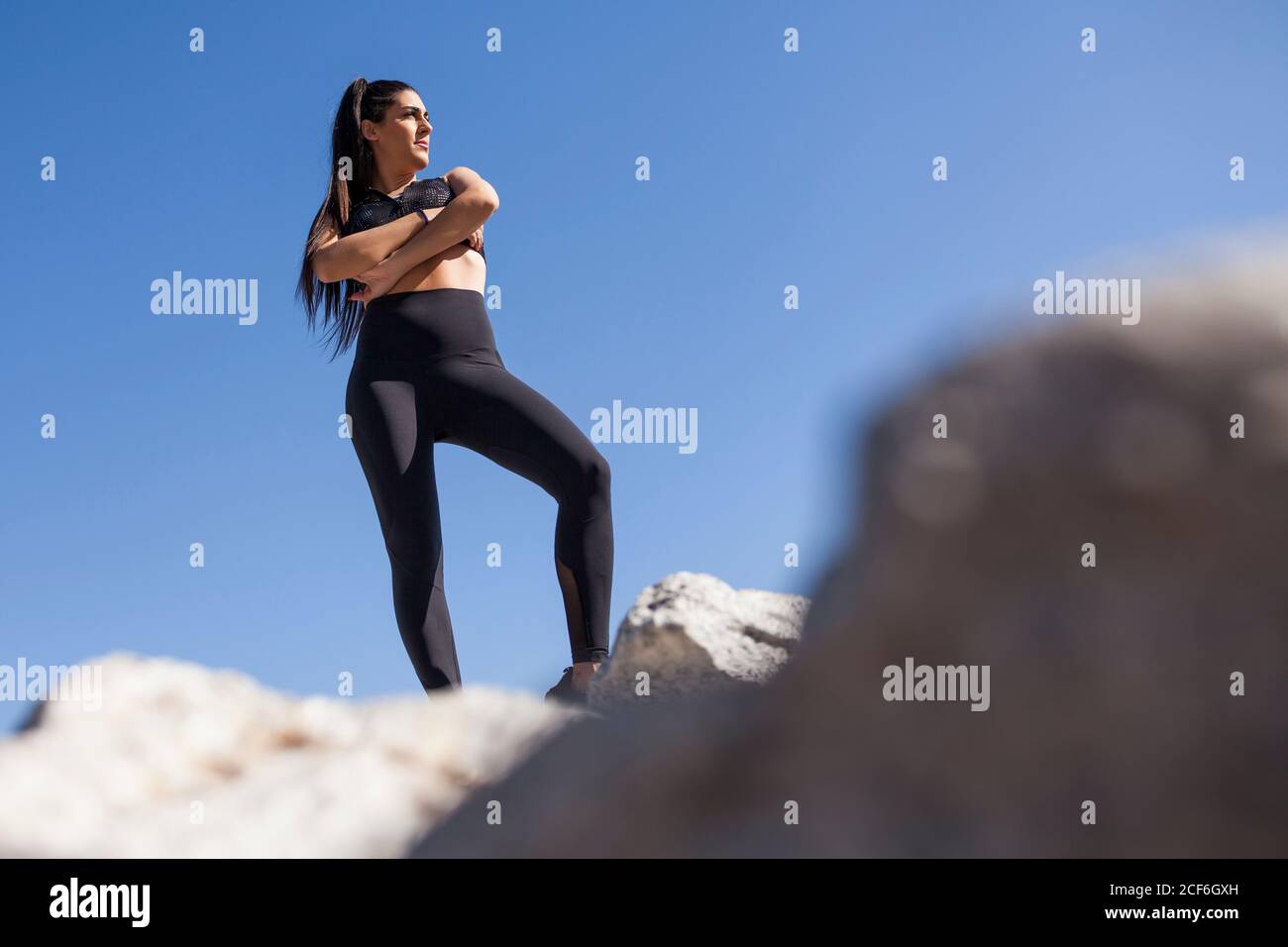 Von unten junge Brünette Frau in Sportkleidung mit gekreuzten Händen posiert auf Felsen in sonnigen Tag Stockfoto