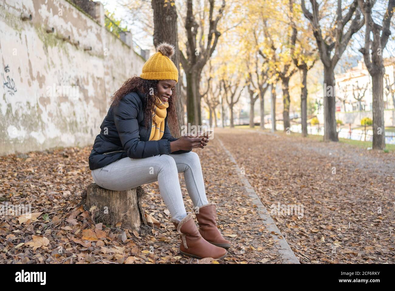 Afroamerikanische Frau in gelbem Hut und warmer Jacke mit Smartphone auf der Straße mit Herbstblättern im Park sitzen Stockfoto