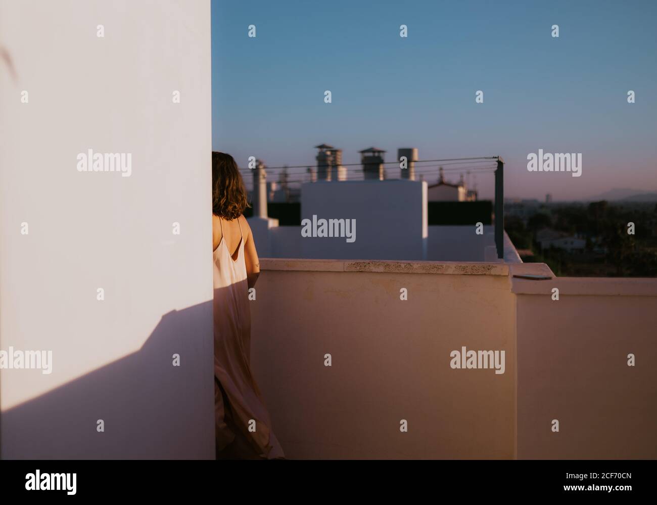 Rückansicht der gesichtslosen Frau in hellrosa Seidenkleid Man konnte um die Ecke auf einem Balkon mit blauem Himmel laufen Und grüne Pflanzen auf dem Hintergrund Stockfoto