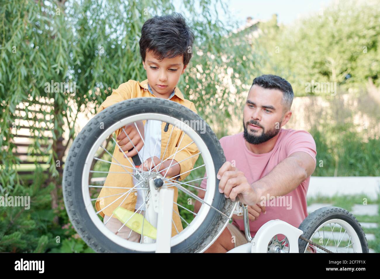 Junger Vater hält Fahrrad, während sein kleiner Sohn versucht, sein Rad zu reparieren, horizontal Medium Schuss Stockfoto