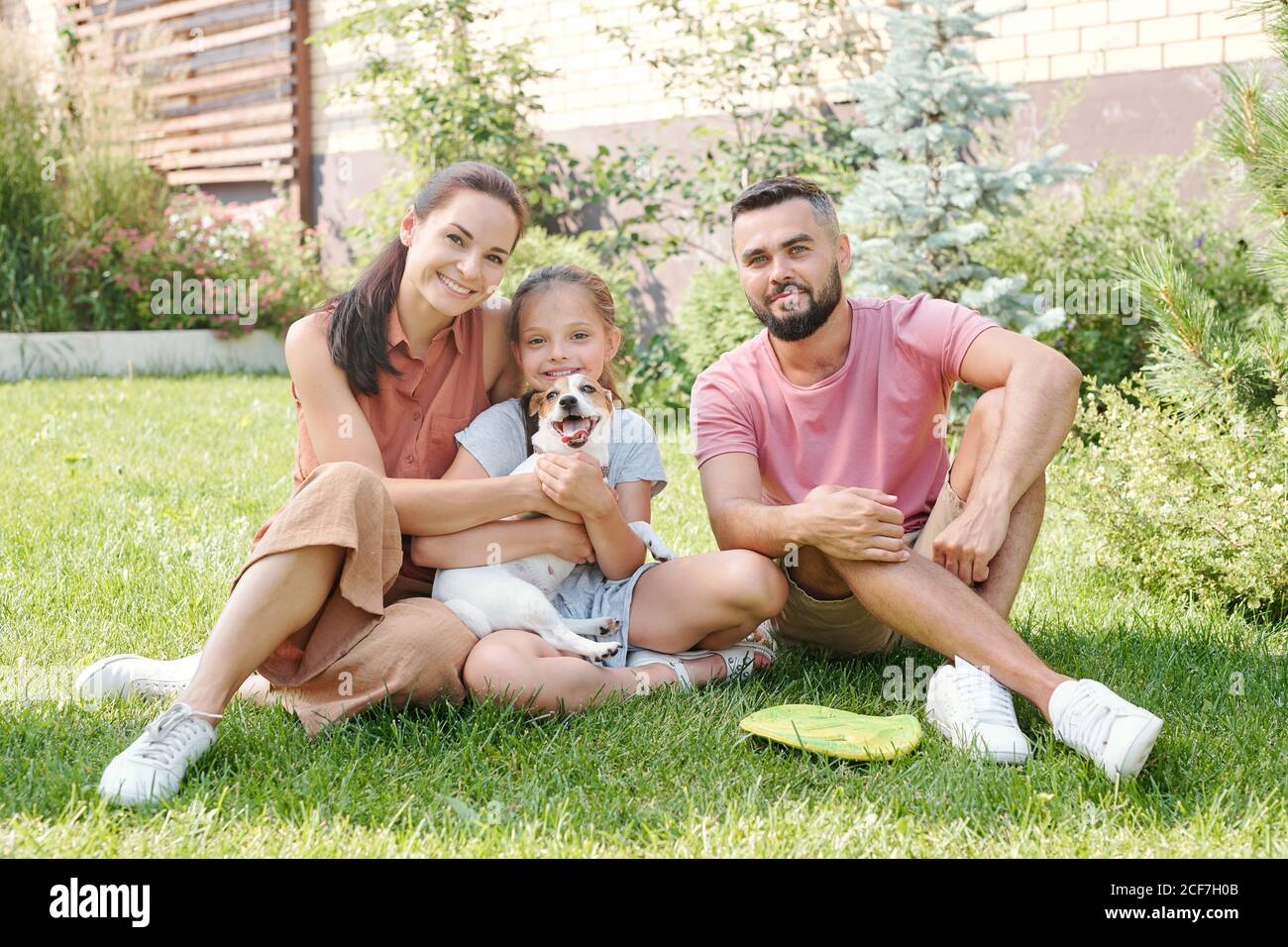 Junge Familie mit einer Tochter verbringen Zeit mit ihren Kleiner Hund im Hinterhof sitzt auf dem Rasen lächelt an der Kamera Stockfoto