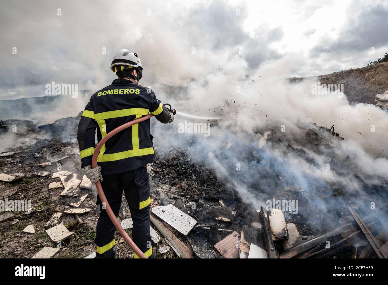 Rückansicht des tapferen Feuerwehrmann in schützender Uniform stehend mit Schlauch und Löschfeuer auf Müllhalde in Bergen Stockfoto