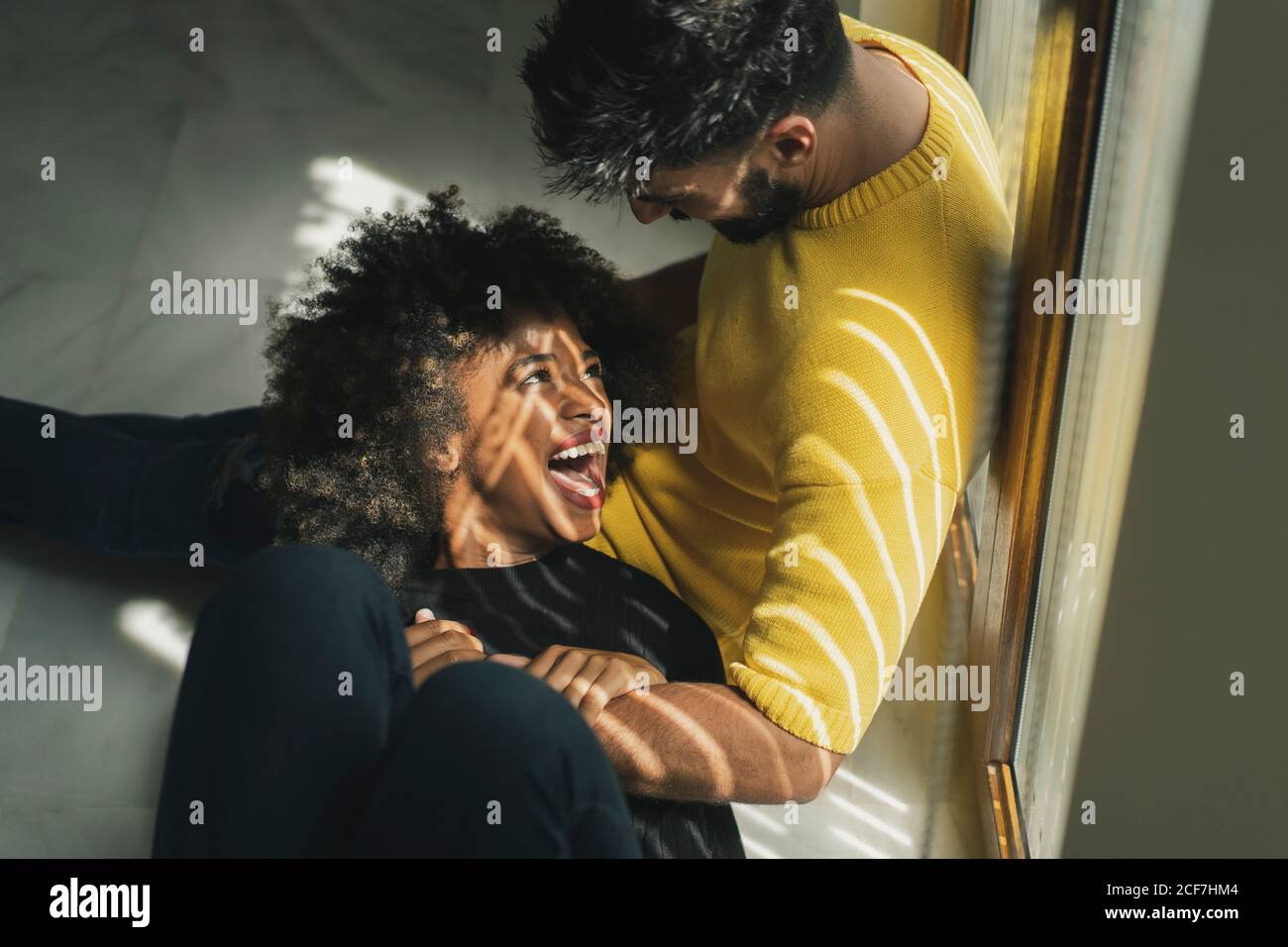 Ziemlich fröhliche afroamerikanische Frau, die lachend auf den Knien eines bärtigen Mannes in einem stilvollen Zimmer lag Stockfoto