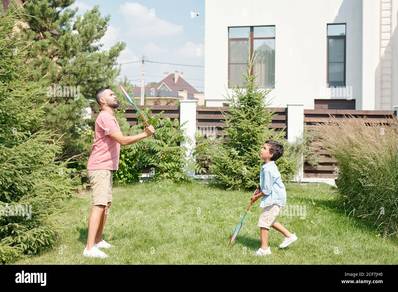Junge Erwachsene Mann in lässigem Outfit Badminton spielen mit seinem kleinen Sohn auf Rasen im Hinterhof, Seitenansicht erschossen Stockfoto