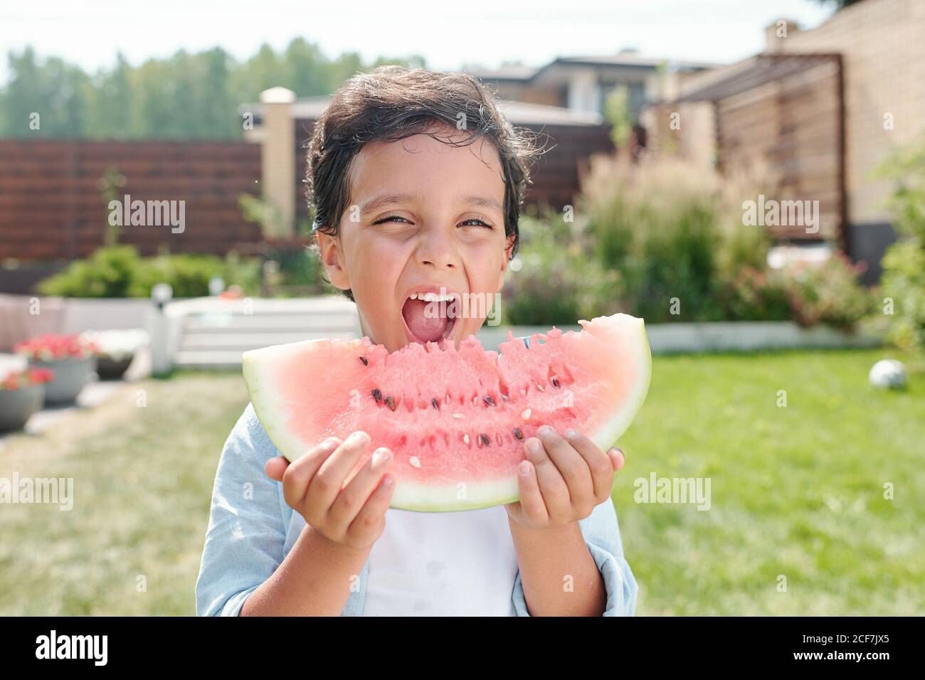 Mittellange Nahaufnahme des fröhlichen kleinen Jungen, der im Bild steht Hinterhof essen Stück Wassermelone Stockfoto
