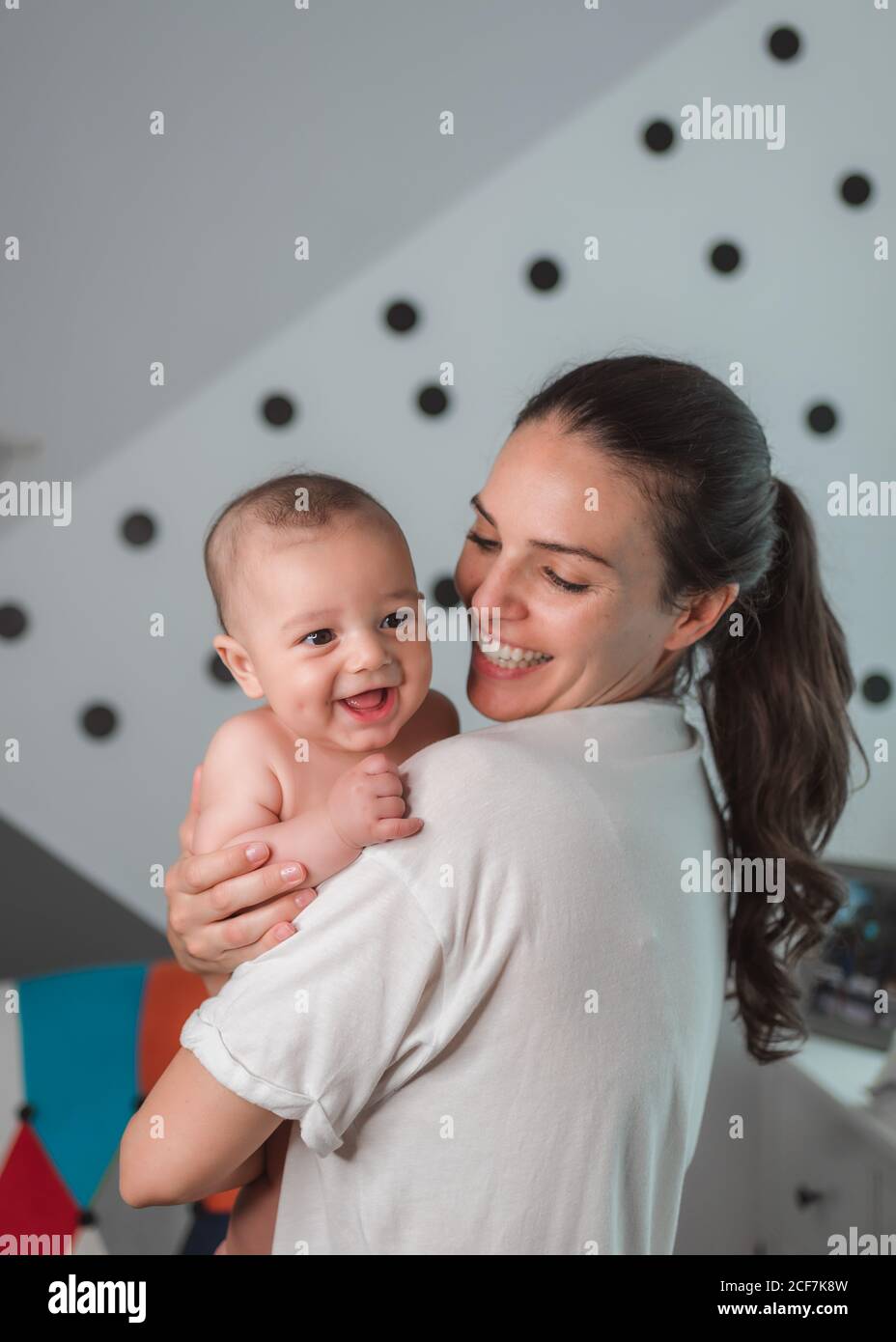 Seitenansicht der zufriedenen jungen Mutter in lässiger Kleidung lachend Mit Inhalt Baby in den Armen während sonnigen Morgen Stockfoto