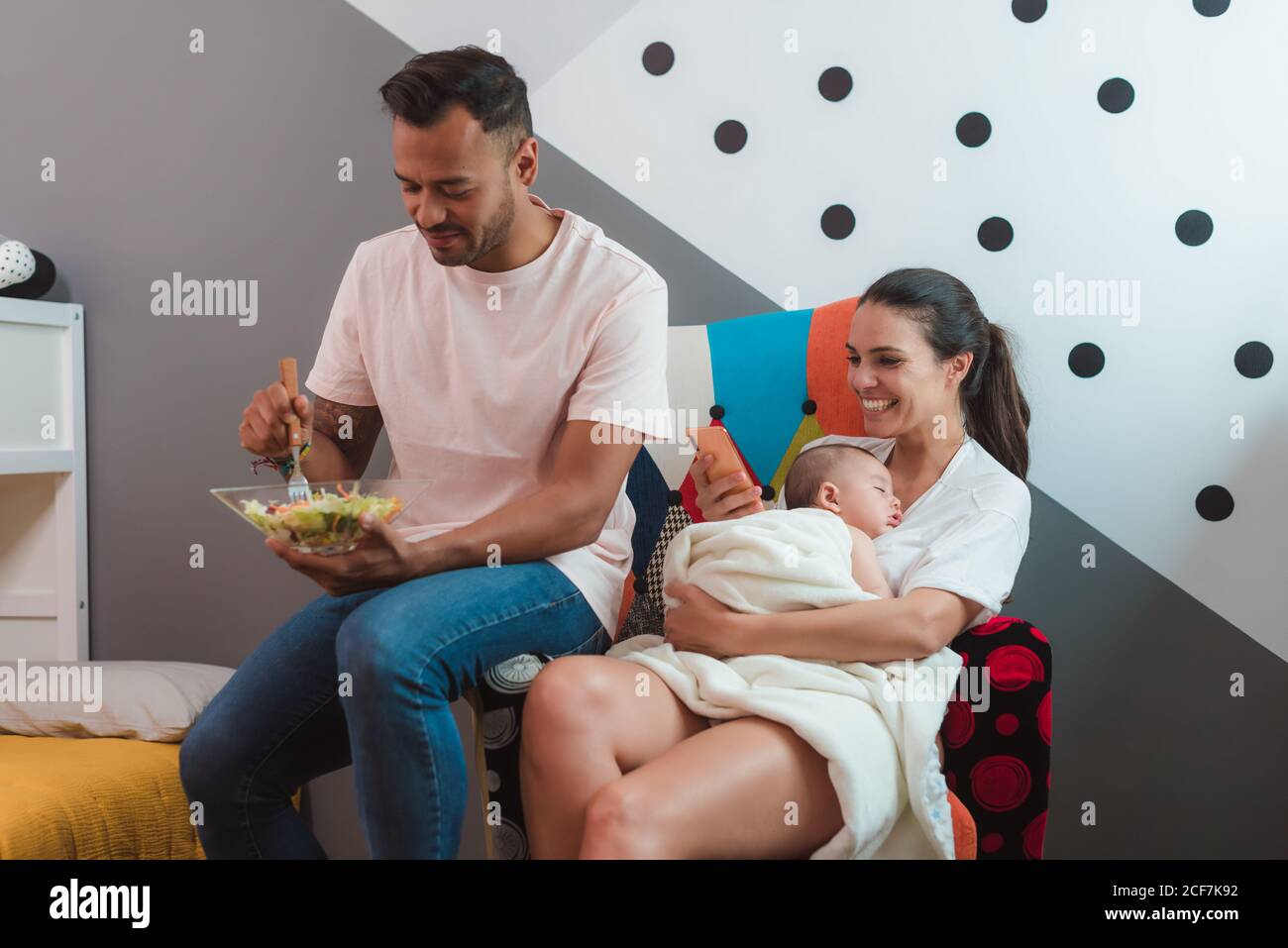 Schwarz haarige Mutter mit schläfrigen Baby ob Brust mit Handy Telefon in der Hand sitzend auf Stuhl mit Vater in der Freizeit Tragen Sie essenden Salat Stockfoto