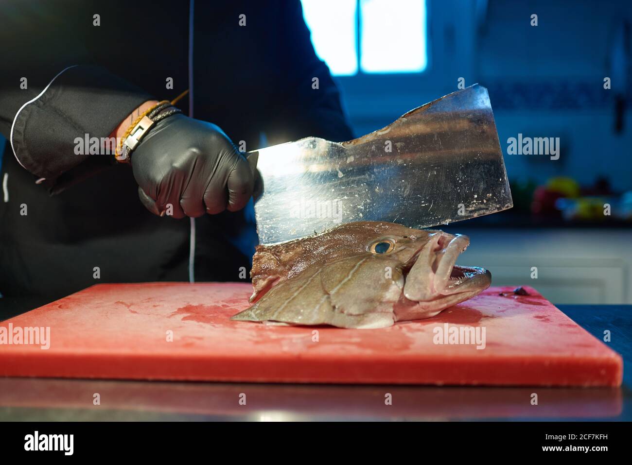 Unkenntlich garen in schwarzer Uniform und Handschuhe mit Metallspalter Beim Schneiden Kopf von großen frischen Fisch auf rot gehackt Tafel am Tisch gegen verschwommenes Interieur der modernen Küche Stockfoto