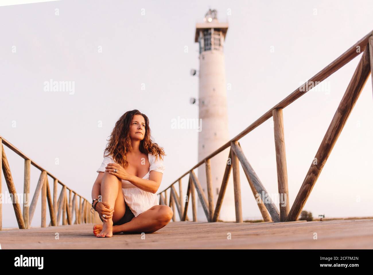 Nachdenkliche gebräunte Frau in sommerlicher offener Kleidung sitzt mit gekreuzten Barfuß-Beinen auf einer Holzbrücke zum Leuchtturm in Fuerteventura, Las Palmas, Spanien Stockfoto