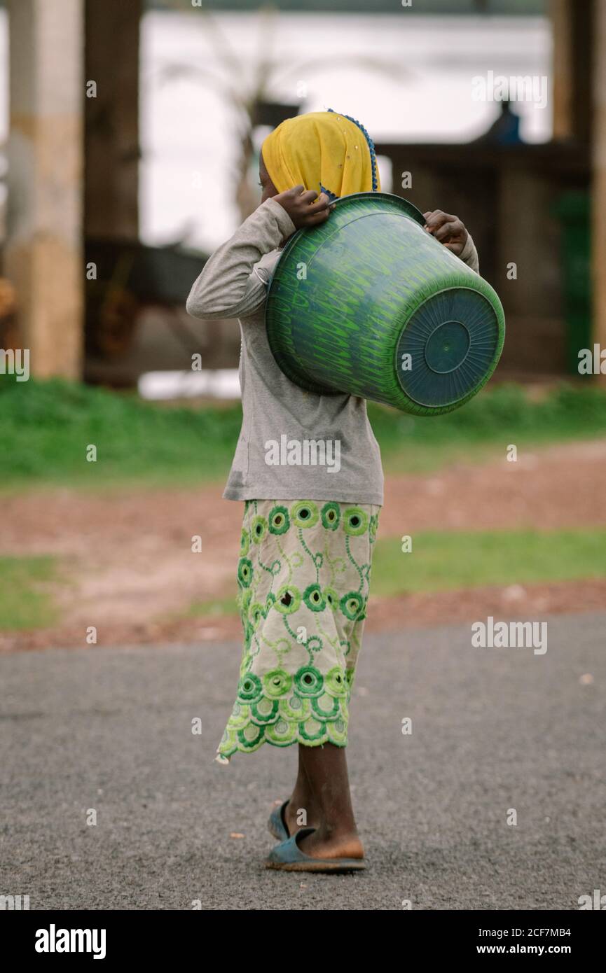 Gambia, Afrika - 7. August 2019: Rückansicht eines kleinen armen Kindes in einfacher Kleidung, das während des Spaziergangs auf der Straße grünes Becken auf der Rückseite hält Stockfoto