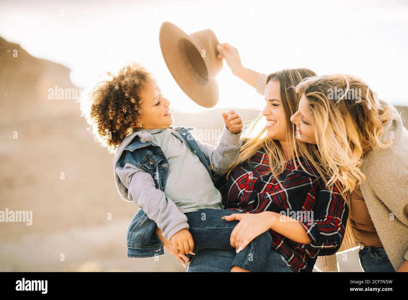 Fröhliche Frau im Karomuster hält in den Armen lässig Kleinkind mit lockigem Haar, während fröhliche Freundin Hut auf Kind auf die Natur am Tag Stockfoto