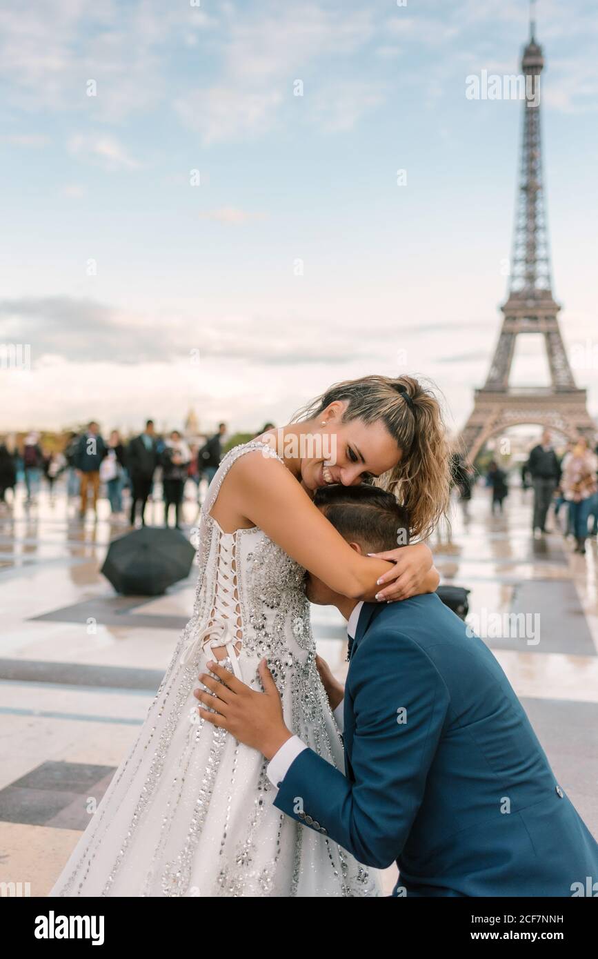Inhalt Bräutigam in blau stilvoller Anzug auf Knie und Küssen zufriedene Braut in weißem Hochzeitskleid mit Eiffelturm Im Hintergrund Stockfoto