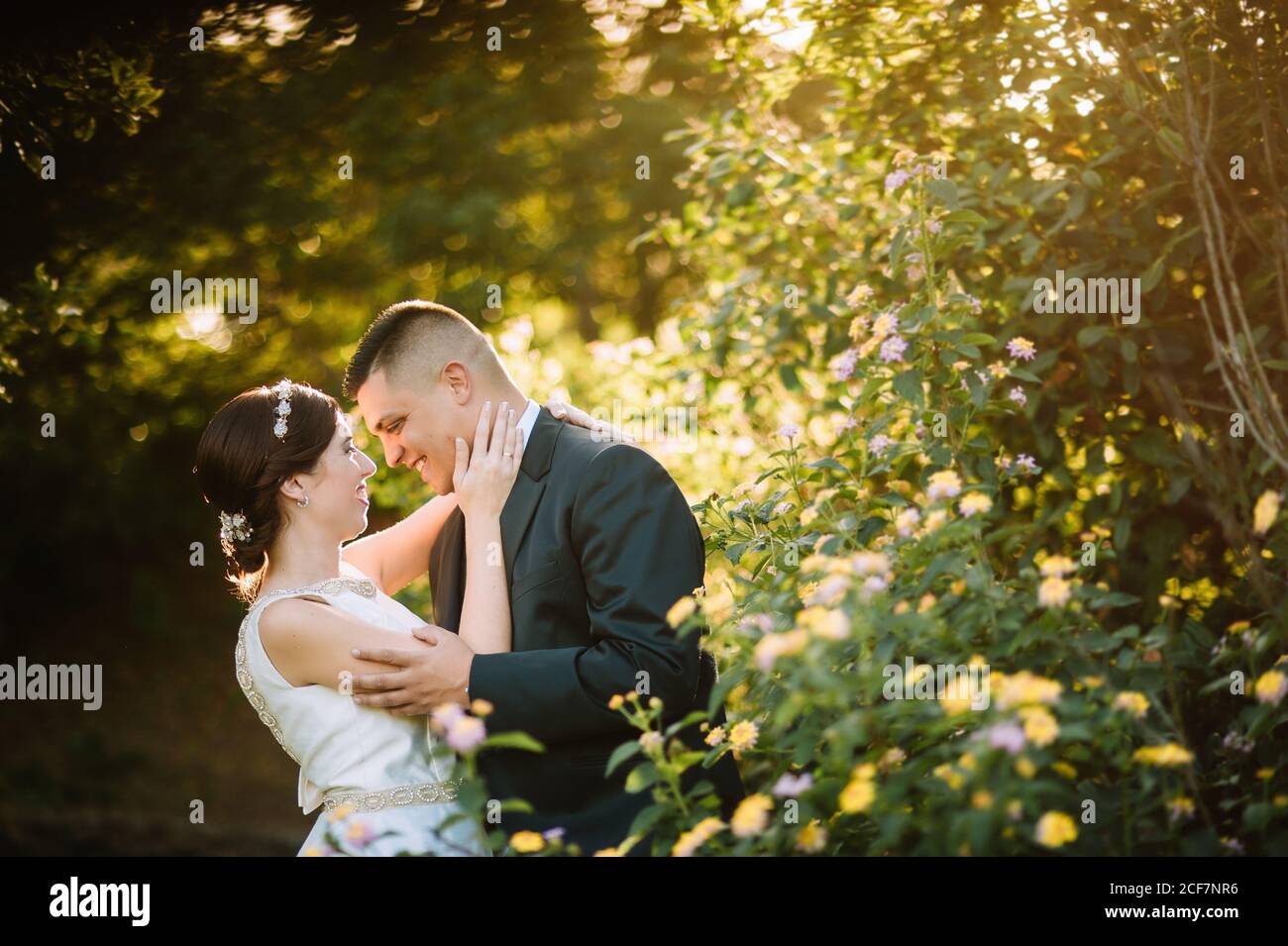 Seitenansicht der anmutigen zarten Brautpaar Bonding und suchen An die Augen mit Liebe zwischen leuchtend gelben Blumen und Büschen Im Garten Stockfoto