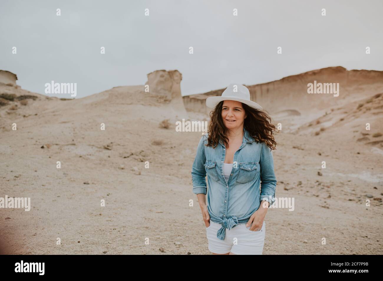 Positive Frau in legerer Kleidung, die einen Hut trägt, mit Händen in den Taschen an sandigen Dünen herumläuft und an einem düsteren Tag wegschaut Stockfoto