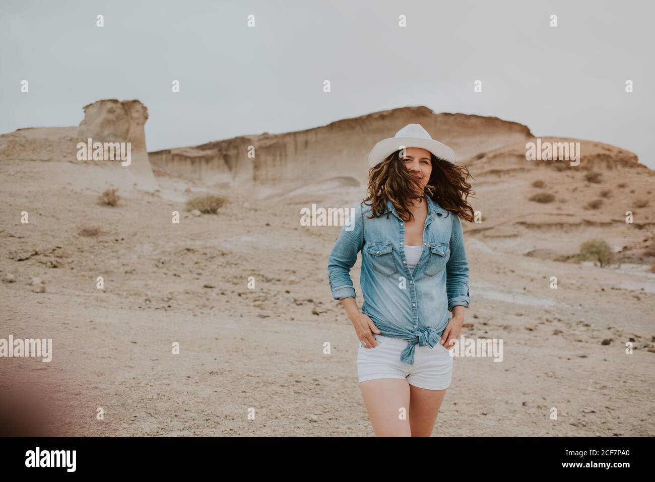 Positive Frau in legerer Kleidung, die einen Hut trägt, mit Händen in den Taschen an sandigen Dünen herumläuft und an einem düsteren Tag wegschaut Stockfoto