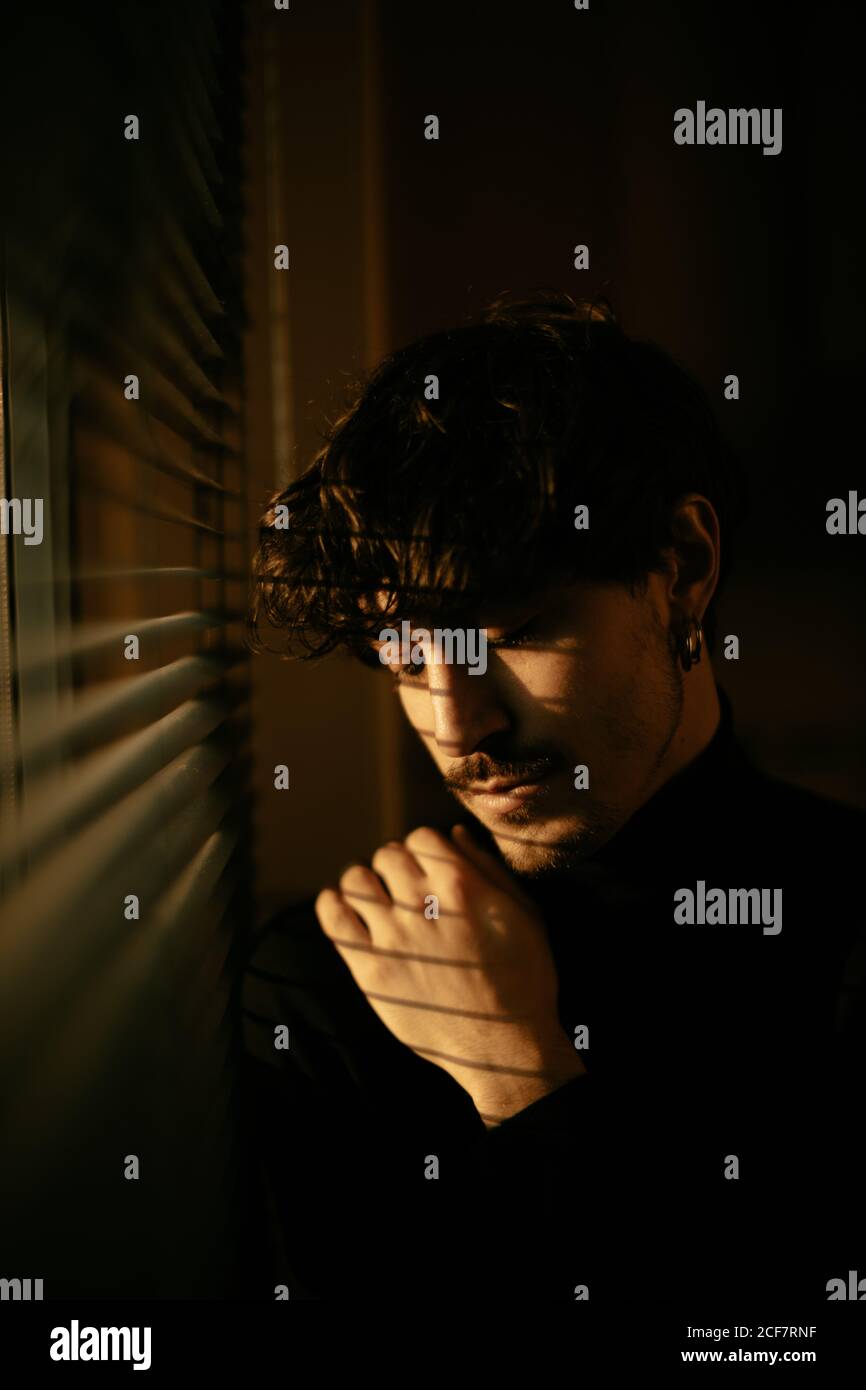 Junger melancholischer Kerl im schwarzen Rollkragen, der neben dem Fenster steht Mit Fensterläden mit Schatten auf Gesicht Stockfoto