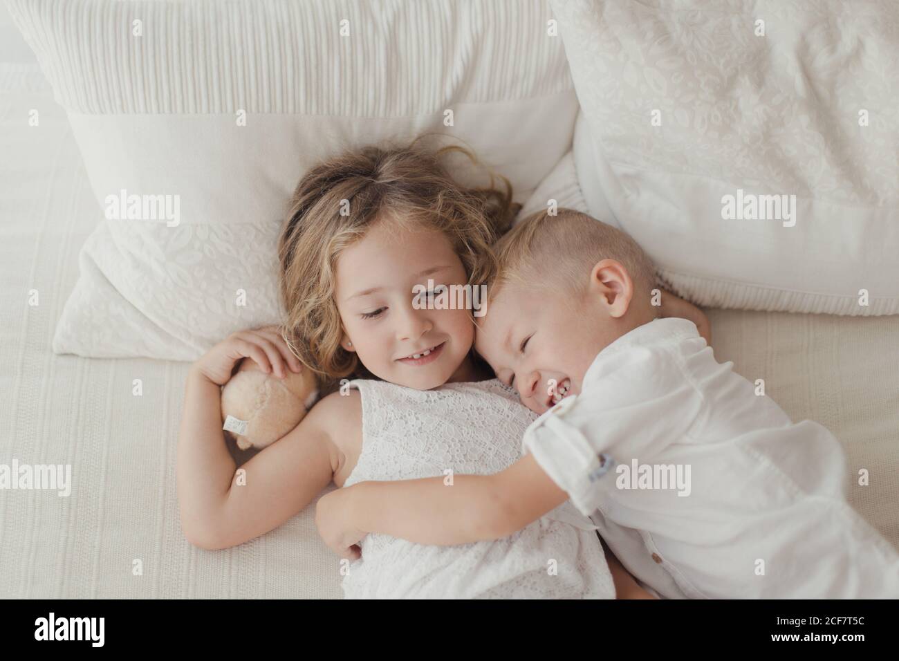 Von oben glückliche männliche und weibliche Kinder in weiß Kleidung liegt auf dem Bett in einer Umarmung und lächelnd Stockfoto