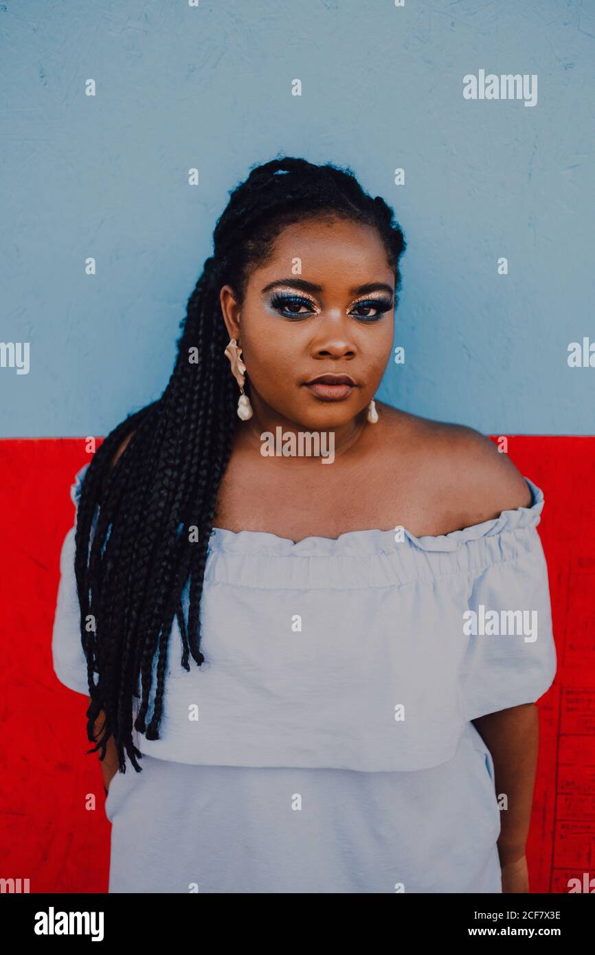 Attraktive moderne junge schwarze junge Frau mit hellen Make-up in Off-Shoulder-Kleid stehend Blick auf die Kamera Stockfoto