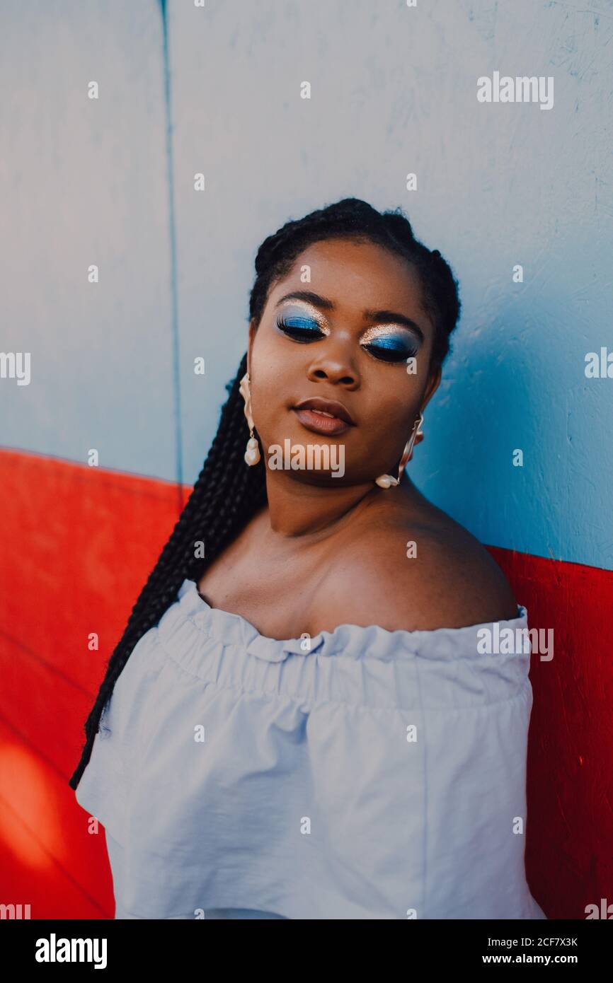 Attraktive moderne junge schwarze junge Frau mit hellen Make-up in Off-Shoulder-Kleid stehend mit geschlossenen Augen Stockfoto