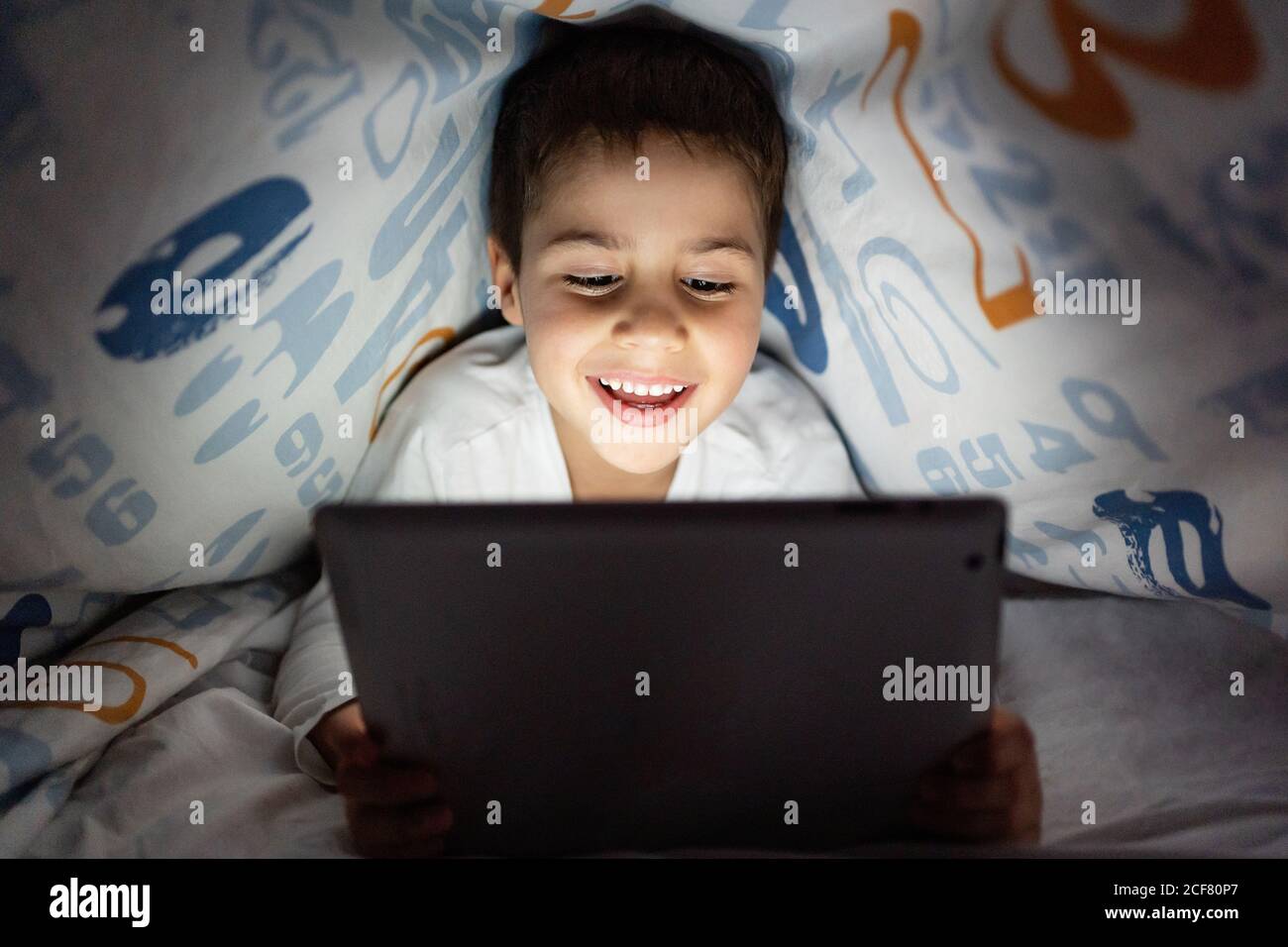 Adorable Kind in Pyjamas versteckt unter Decke und mit Tablet Während der Unterhaltung während der Nacht zu Hause Stockfoto