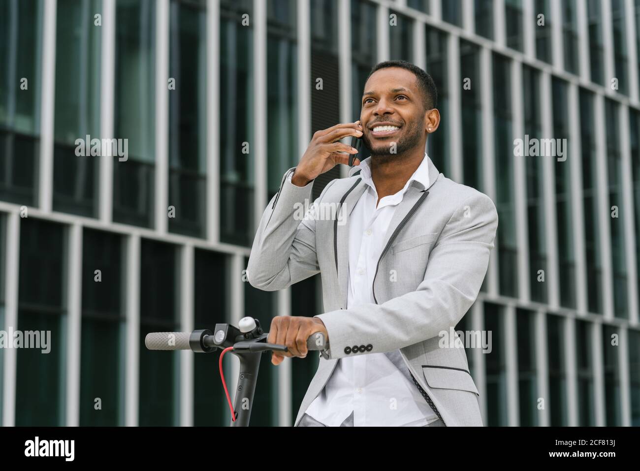 Elegante afroamerikanische fröhliche Geschäftsmann Reiten Roller und im Gespräch Handy in der Innenstadt Stockfoto