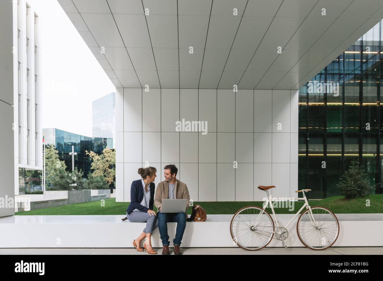 Entzückende Geschäftsleute, die lächelten und gemeinsam mit dem Laptop unterwegs waren Modernes Gebäude in der Nähe des Fahrrads an der Stadtstraße Stockfoto