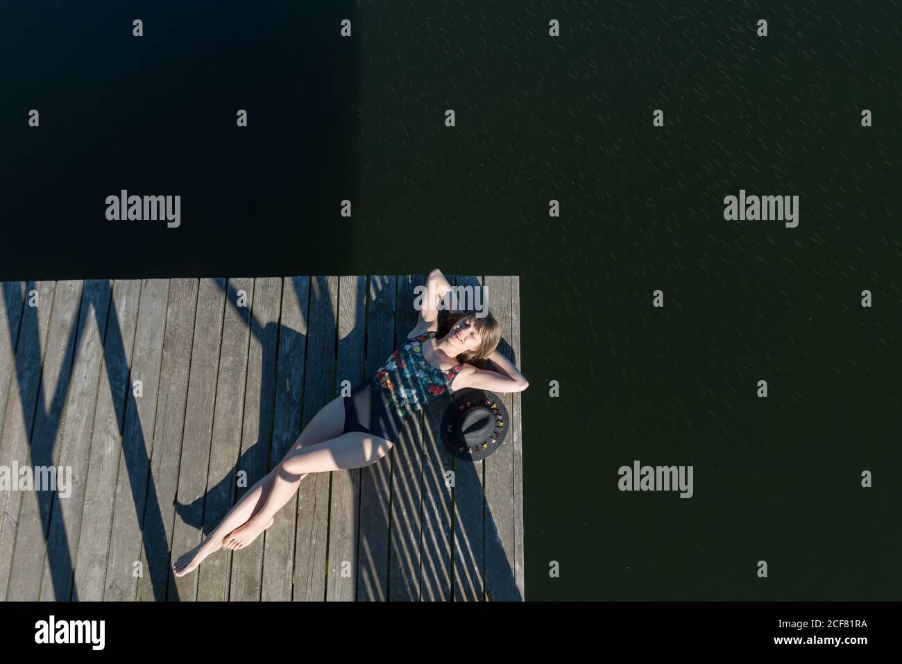 Draufsicht auf eine wunderschöne Frau in schwarzem Badeanzug und Hut, die auf dem hölzernen Pier des Sees auf dem klaren blauen Himmel und im Waldhintergrund liegt Stockfoto