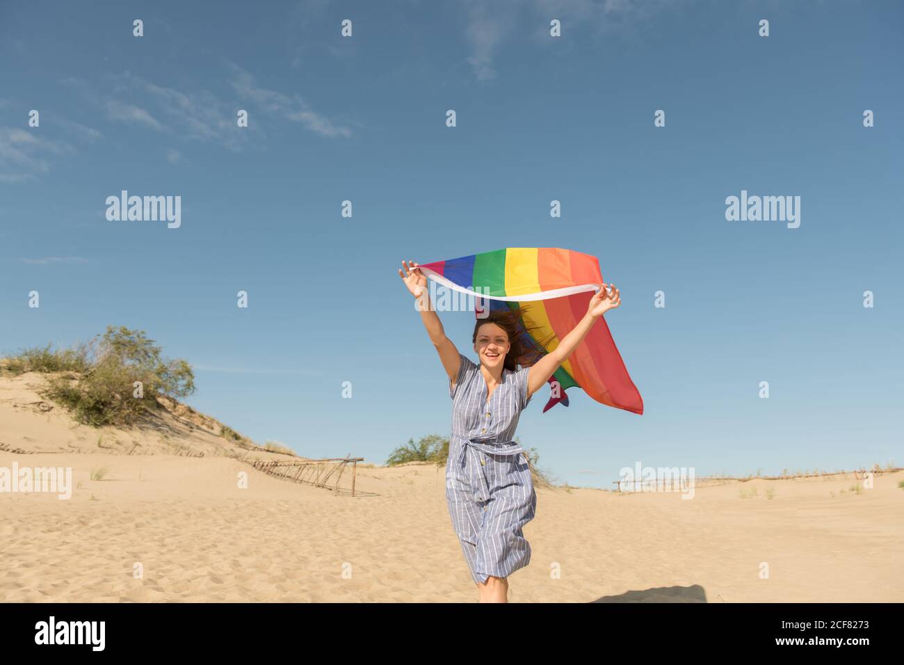 Erwachsene selbstbewusste Frau in legerem Kleid, die an windigen Tagen eine Regenbogenflagge über dem Kopf auf Sanddünen trägt Stockfoto