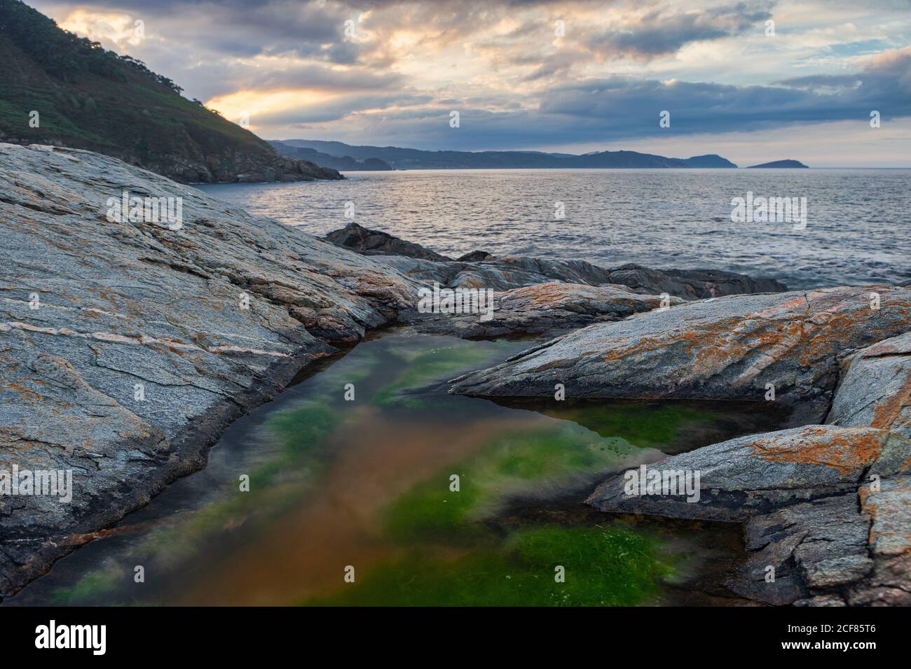 Von oben ein kleines Pfund mit Moos zwischen mächtigen Felsen Mit ruhigen Meer überwuchert Berge und wolkigen Himmel auf Hintergrund Am Portonovo Strand Stockfoto