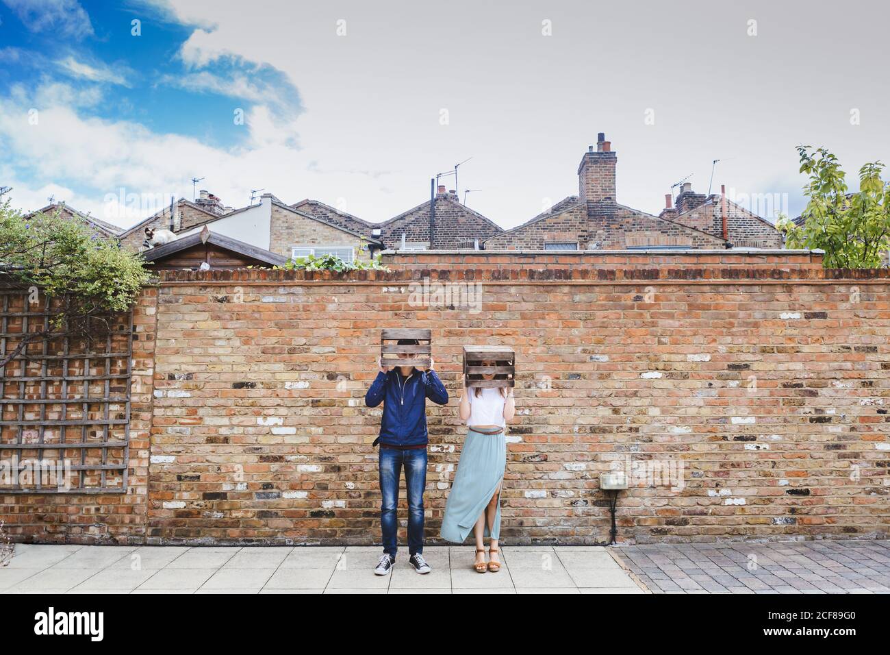 Paar in legerer Kleidung, die in der Nähe einer Ziegelmauer in der Stadt steht Und beim Verhalten hölzerne Obstkisten auf den Köpfen aufsetzen Lächerlich Stockfoto