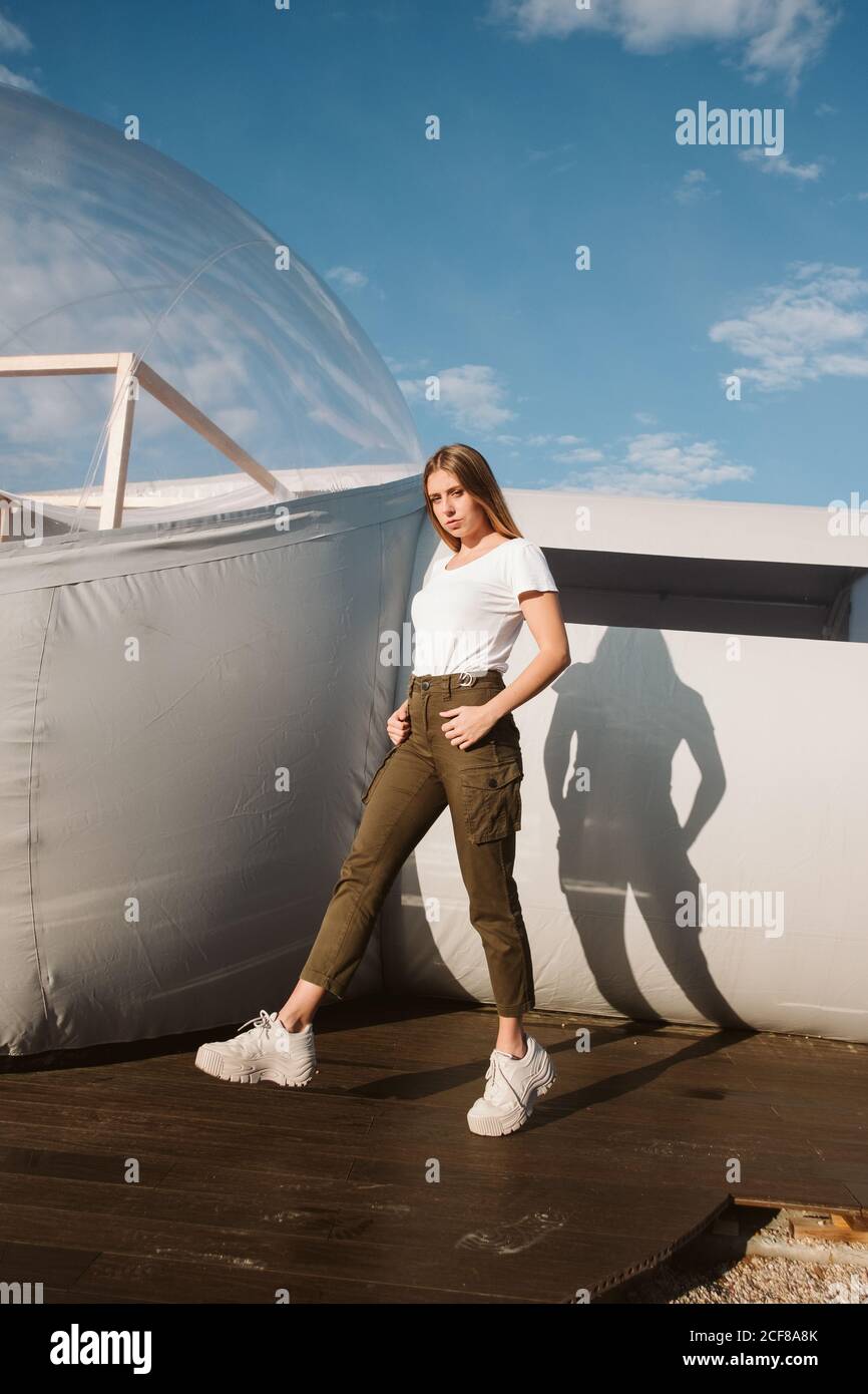 Junge schöne Frau in weißem T-Shirt, Hosen und Sneakers posiert durch transparentes Glamping auf blauem Himmel Hintergrund Stockfoto
