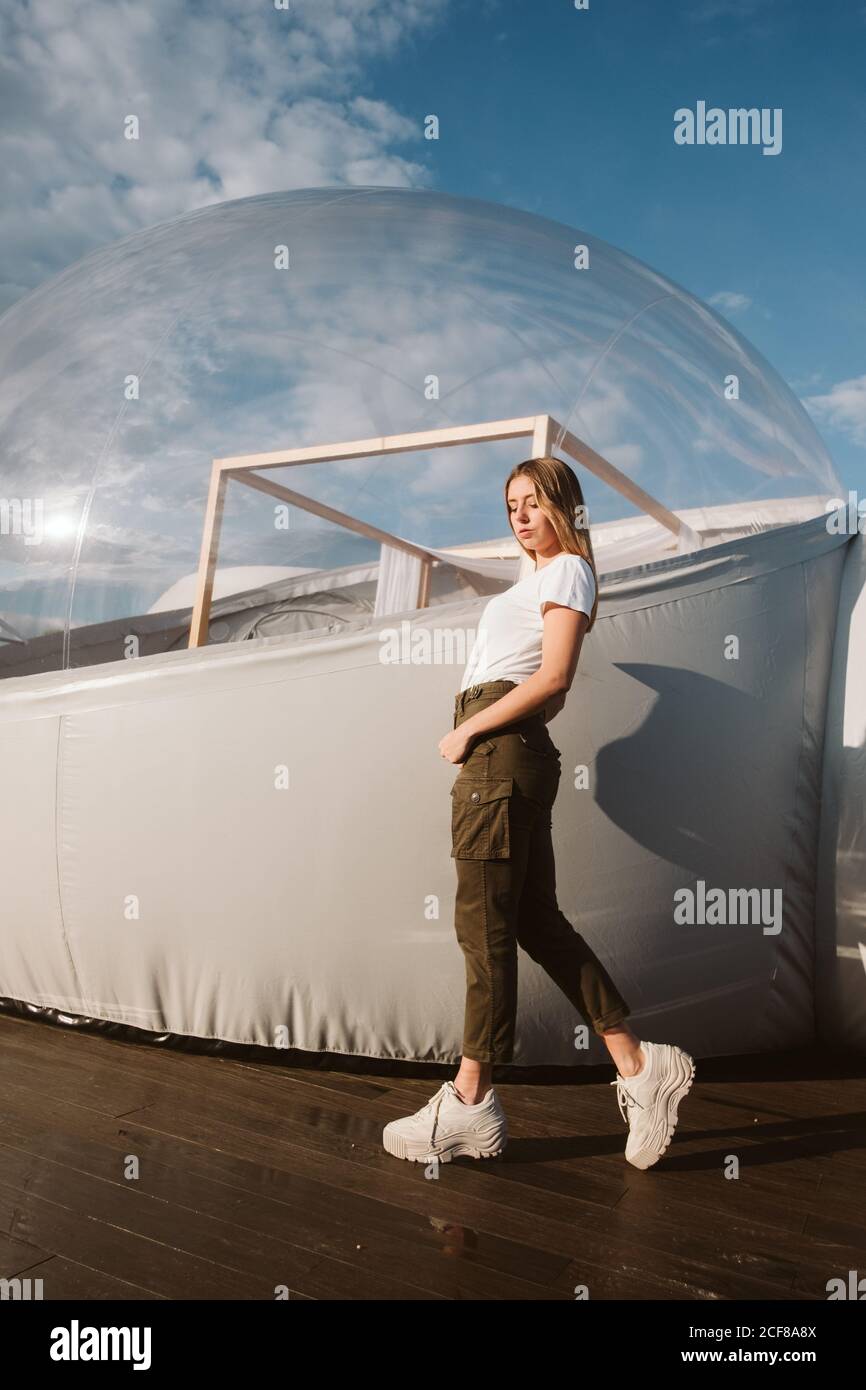 Junge schöne Frau in weißem T-Shirt, Hosen und Sneakers posiert durch transparentes Glamping auf blauem Himmel Hintergrund Stockfoto