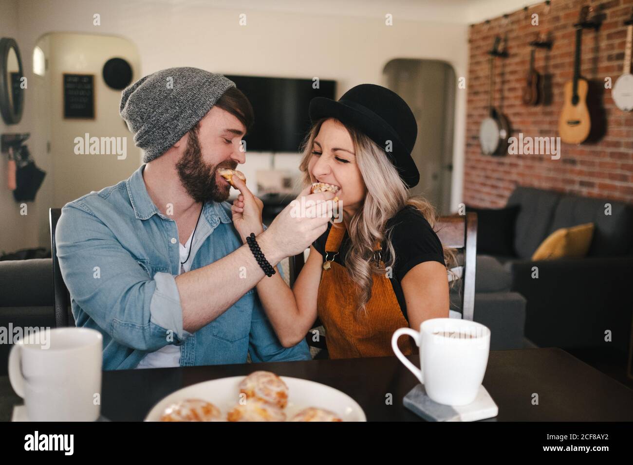 Glückliche Partner in legerer Kleidung füttern sich gegenseitig mit süß Rollt, während die Augen geschlossen werden und am Tisch mit Tassen sitzt Von Kaffee in gemütlichen Wohnung Stockfoto