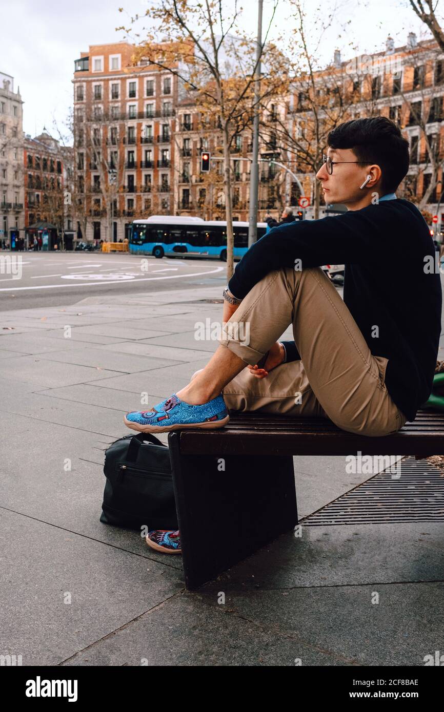 Ganzer Körper von asiatischen jungen trendigen männlichen in stilvollem Outfit Und Brillen sitzen auf kleinen Steinsäule posiert und schaut Entfernt in der Nähe eines alten, wunderschönen Gebäudes an der Straße Stockfoto