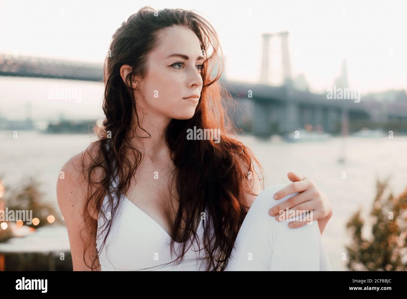 Wunderschöne Frau mit langen Haaren trägt helle Kleidung sitzend auf Hintergrund der Brücke und Fluss während der Entspannung und Blick weg Stockfoto