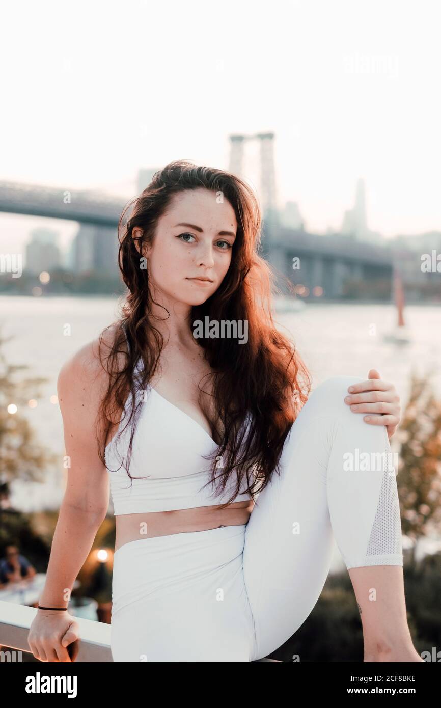 Wunderschöne Frau mit langen Haaren trägt helle Kleidung sitzend auf Hintergrund der Brücke und Fluss während der Entspannung und Blick weg Stockfoto