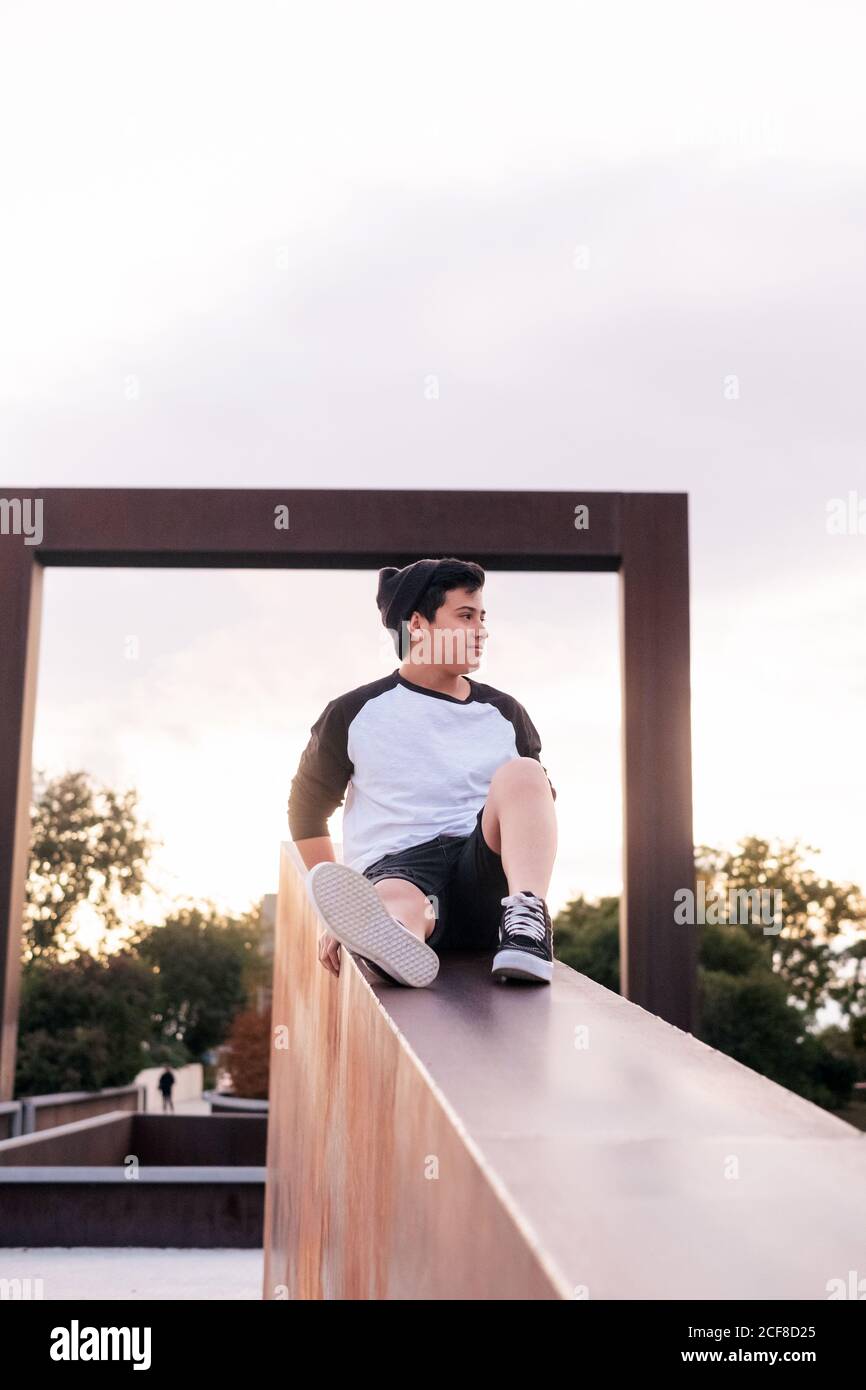 Niedriger Winkel der positiven jungen Mann in Casual tragen sitzen Auf Steinzaun und wegschauen, während Sie sich in der Stadt ausruhen park in sonnigen Abend Stockfoto
