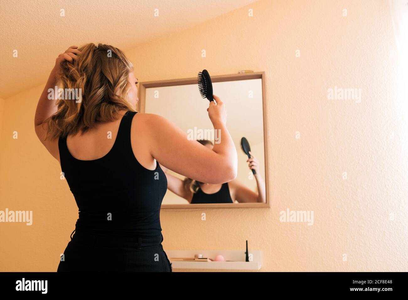 Niedrige Winkel Rückansicht der jungen Frau in lässigem Outfit Haare putzen und Pferdeschwanz machen, während Sie vor stehen Spiegel zu Hause Stockfoto