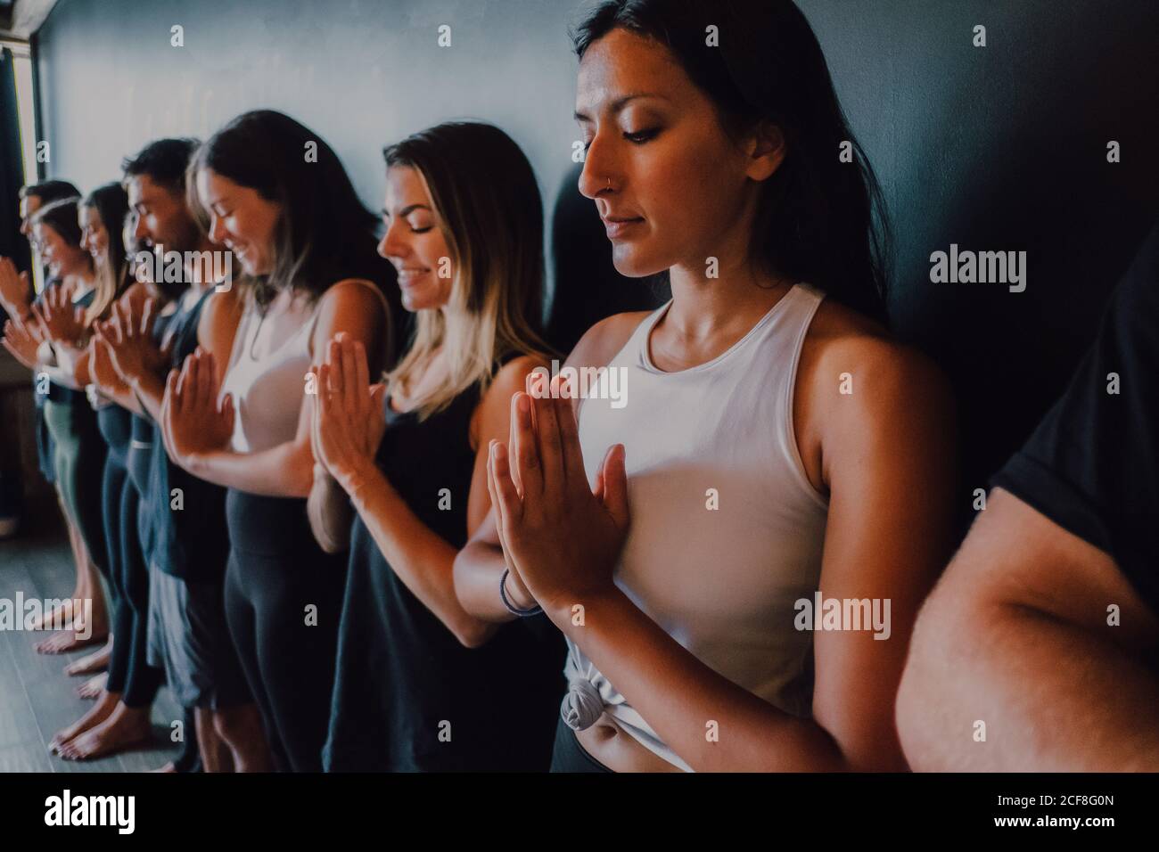 Gruppe von glücklichen jungen sportlichen Menschen in aktiven mit Augen Geschlossen entspannend und tiefes Atmen, während man mit Namaste im Stehen steht tadasana und meditieren neben schwarzer Wand im modernen Yoga-Studio Stockfoto