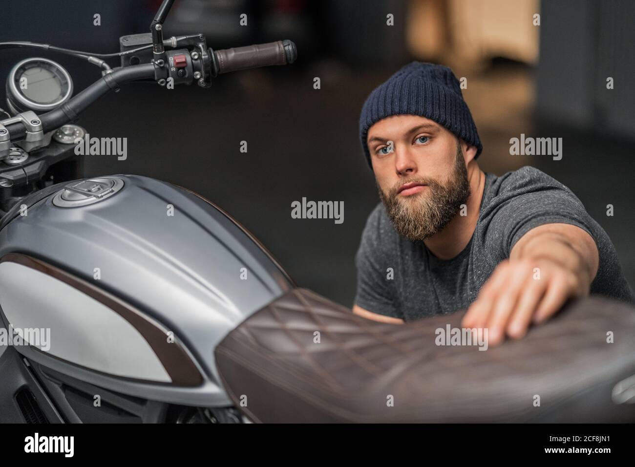 Bärtiger Mann im Hut hob Augenbraue und Blick auf die Kamera Beim Sitzen in der Nähe von Motorrad auf verschwommenem Hintergrund der zeitgenössischen Garage Stockfoto