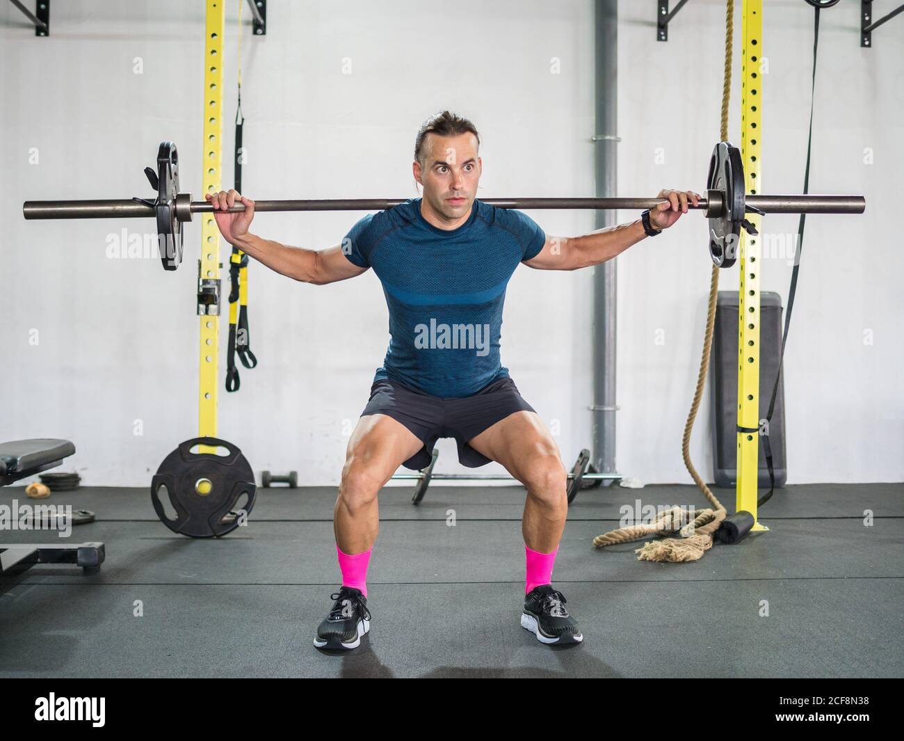 Crouched ruhigen erwachsenen Athleten in Sportkleidung mit Langhantel trainieren Und mit Blick auf weiße Wand und Sportgeräte Sportclub Stockfoto