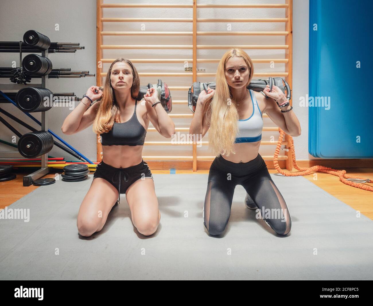 Starke junge Frauen in Sportbekleidung mit schweren Taschen auf den Schultern Und sich gegenseitig angußend, während auf der Turnhalle Matte Gemeinsam Stockfoto